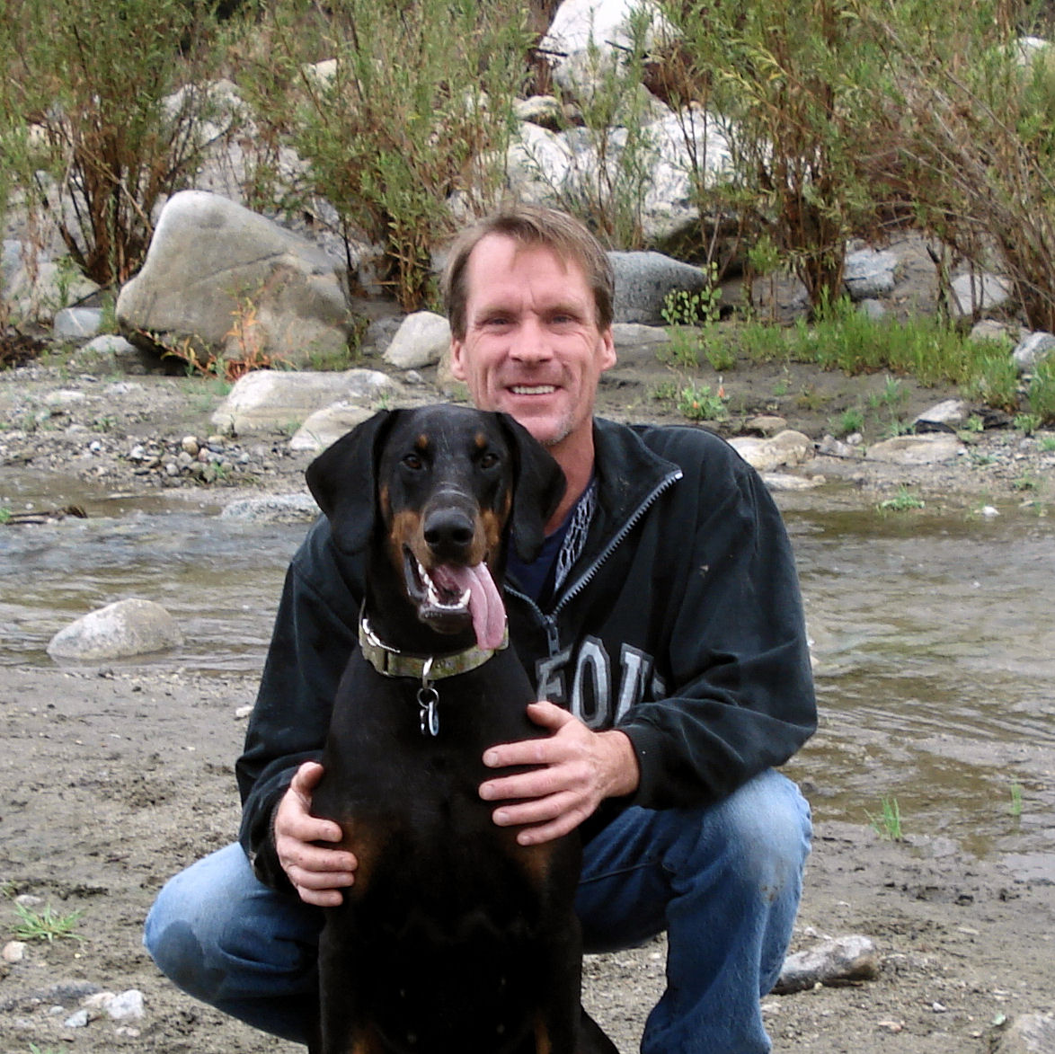 Dave and Chloe. Brown-haired man in mid-50s squats in front of a creek with a doberman whose tongue is hanging out.
